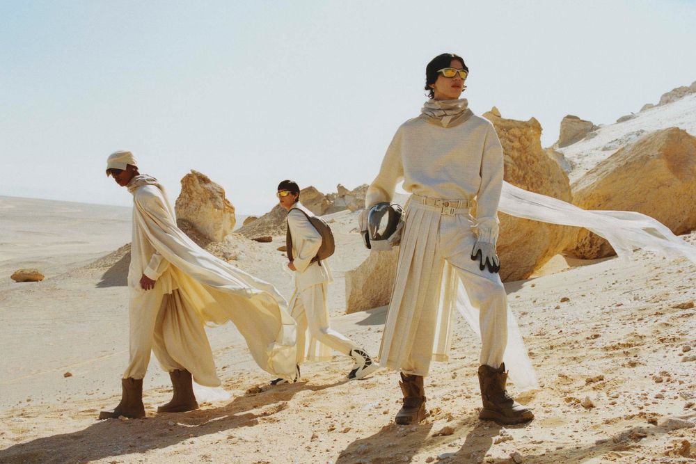 Three people in light-colored, futuristic clothing walk across a desert landscape with rock formations under a clear, bright sky.
