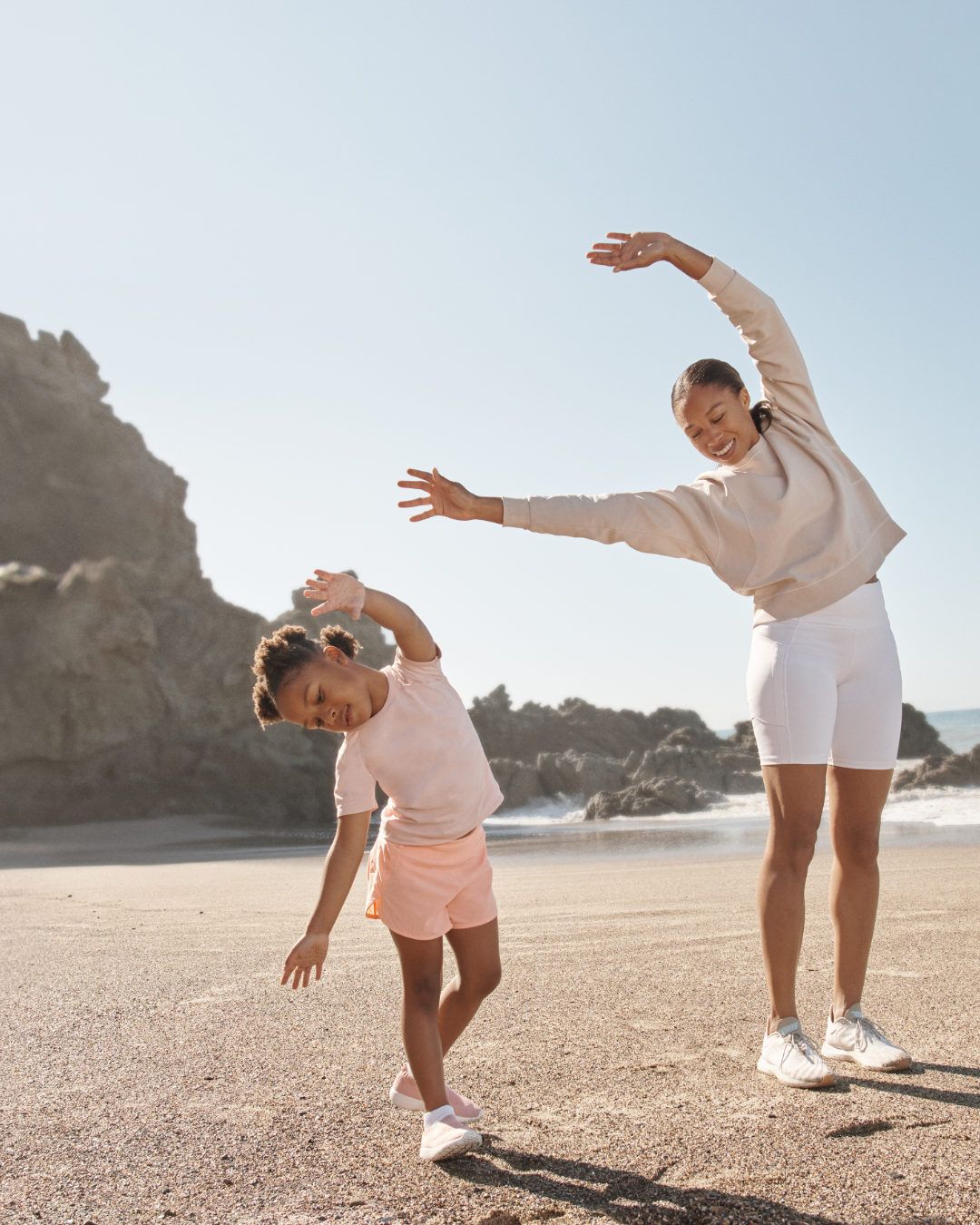 An adult woman and a young girl stretch on a sandy beach next to a rock formation with a clear sky in the background.