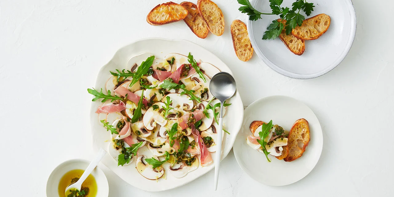 Overhead shot of a mushroom carpaccio dish with prosciutto, arugula, and pesto, served with toasted bread on a white surface.