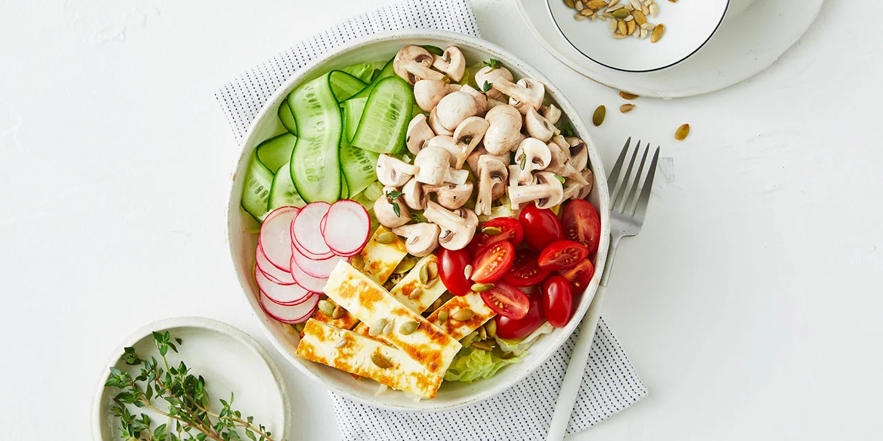 Overhead shot of a vibrant salad bowl with sliced cucumbers, mushrooms, radishes, tomatoes, and grilled cheese, presented on a white table.