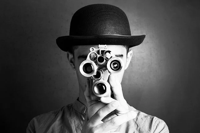 Black-and-white portrait of a man with bowler hat holding a vintage camera obscuring his face, set against a dark gray background.