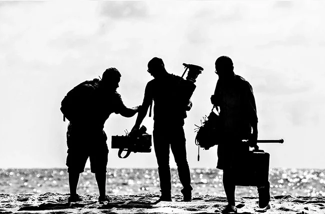 Three silhouetted individuals stand on a beach with camera equipment and luggage, against a bright sky and shimmering ocean backdrop.