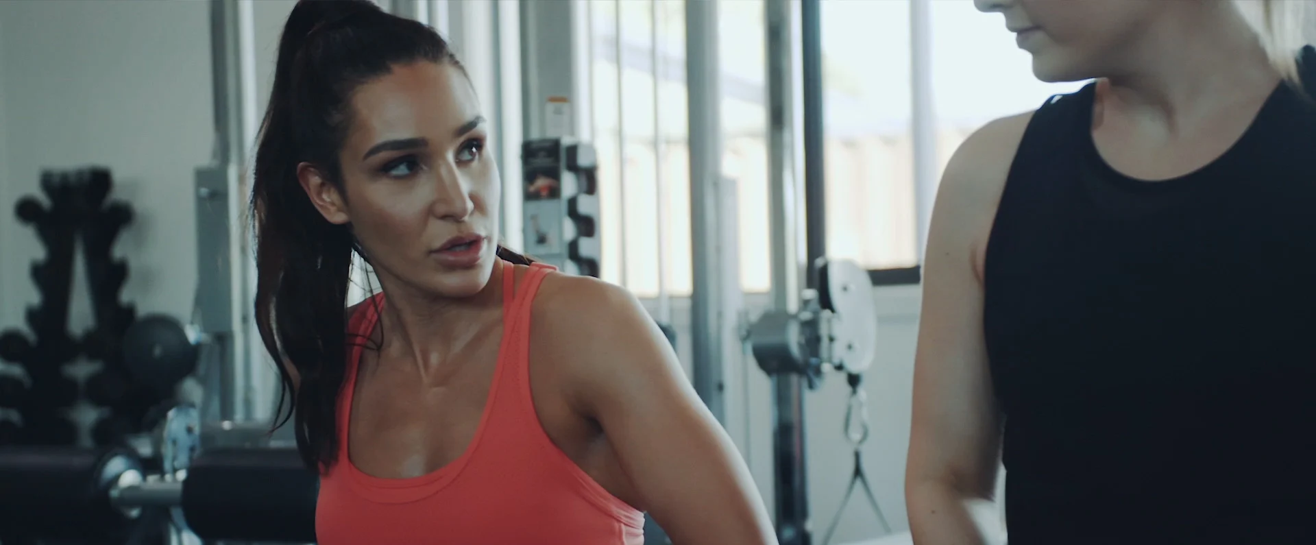 Two women in workout attire are standing in a gym with exercise equipment visible in the background, suggesting a fitness or workout scene.