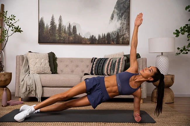 A woman in athletic wear performs a side plank exercise on a mat in a living room with a sofa, artwork, and houseplants.