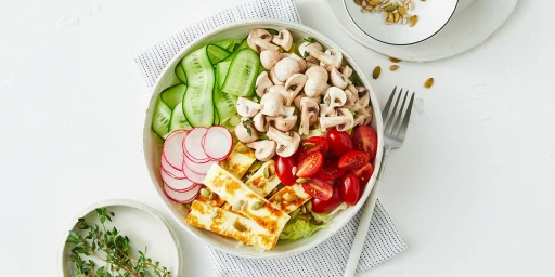 Overhead food photography of a healthy salad bowl with sliced cucumbers, mushrooms, cherry tomatoes, radish, and grilled halloumi cheese.