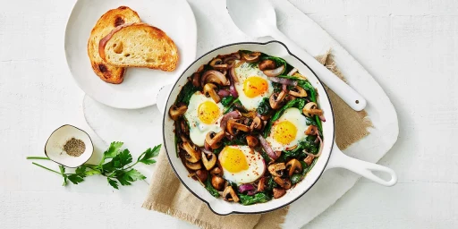 Baked eggs with mushrooms, spinach, and red onion in a white skillet with toasted bread on a white wooden table.