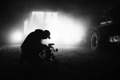 Silhouetted camera operator kneeling next to a camera setup with bright backlit stage lighting and smoke