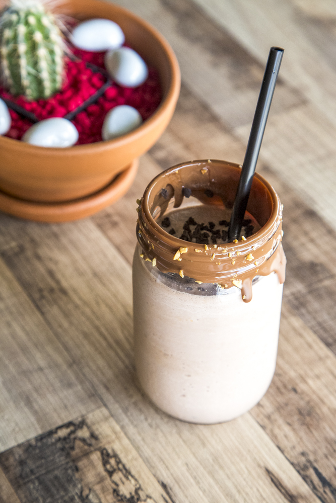 Close-up of a chocolate milkshake in a mason jar with a black straw, next to a cactus in a clay pot on a wooden table.