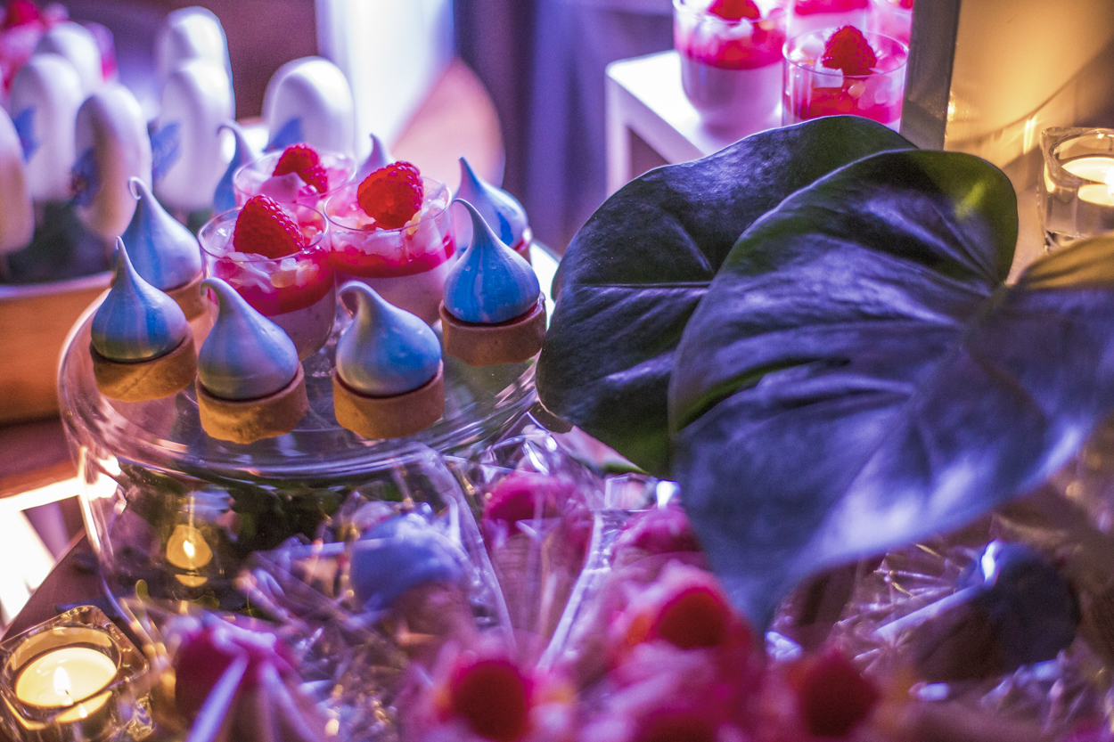 Assorted desserts including meringue drops and raspberry parfaits on a tiered glass stand, with blurred plant in the foreground.