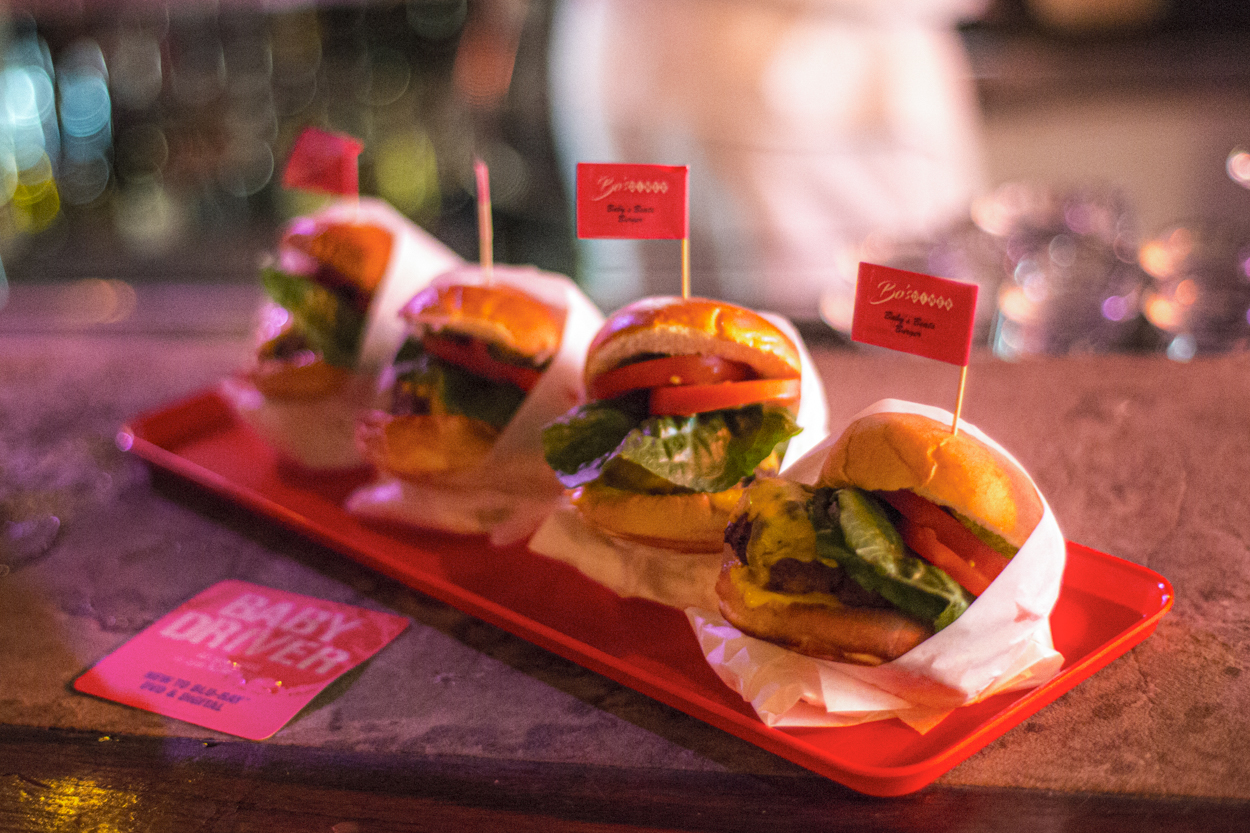 Close-up of four mini-burgers displayed on a red tray, each burger topped with a tiny branded flag, set against a blurred backdrop.