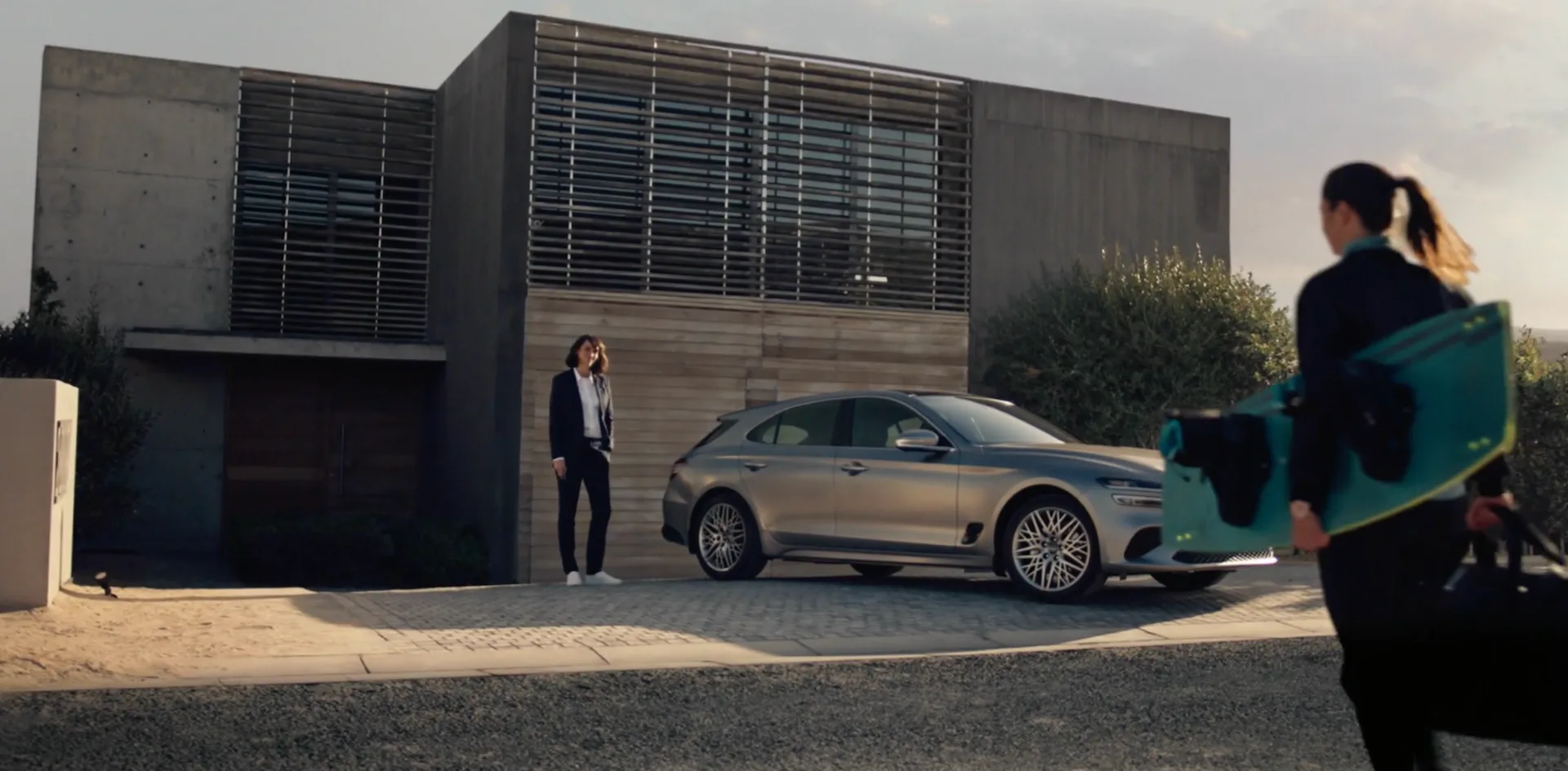 Modern house exterior with silver SUV parked outside; adult woman in business attire stands nearby, another carries a surfboard.