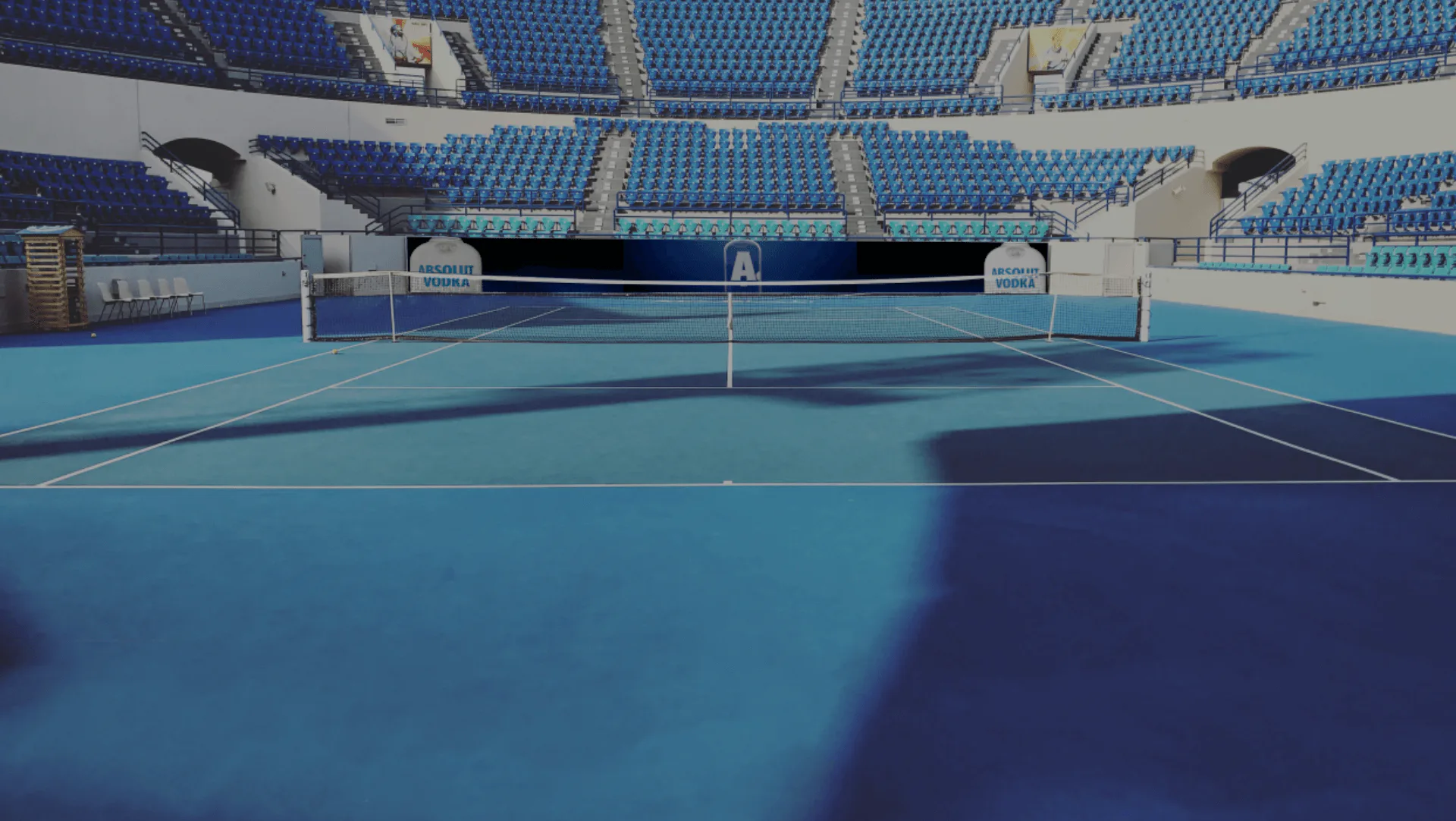 Empty blue tennis court with stadium seating and 'Absolut Vodka' signage. No people are present, but the scene is well-lit.