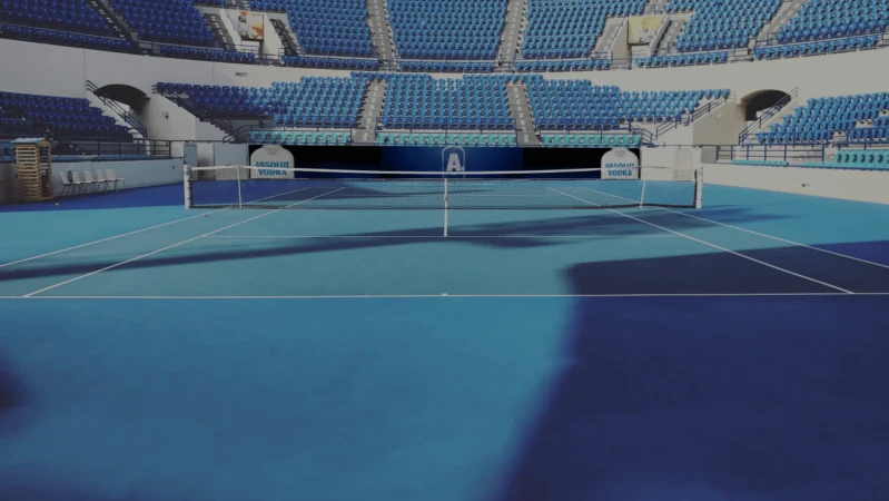 Empty blue tennis court with stadium seating and 'Absolut Vodka' signage. No people are present, but the scene is well-lit.