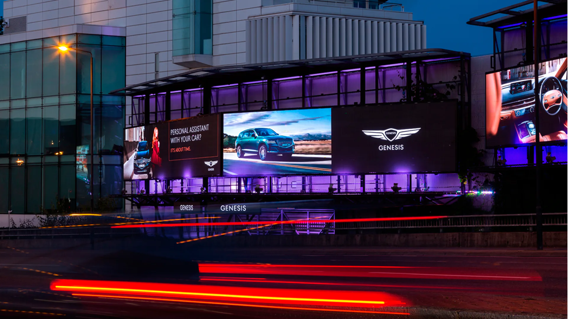 Illuminated Genesis billboards display luxury cars at night with light trails in an urban area. Automotive advertisement showcases car models.
