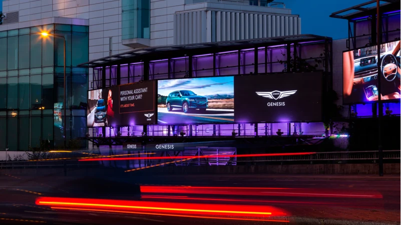 Illuminated Genesis billboards display luxury cars at night with light trails in an urban area. Automotive advertisement showcases car models.