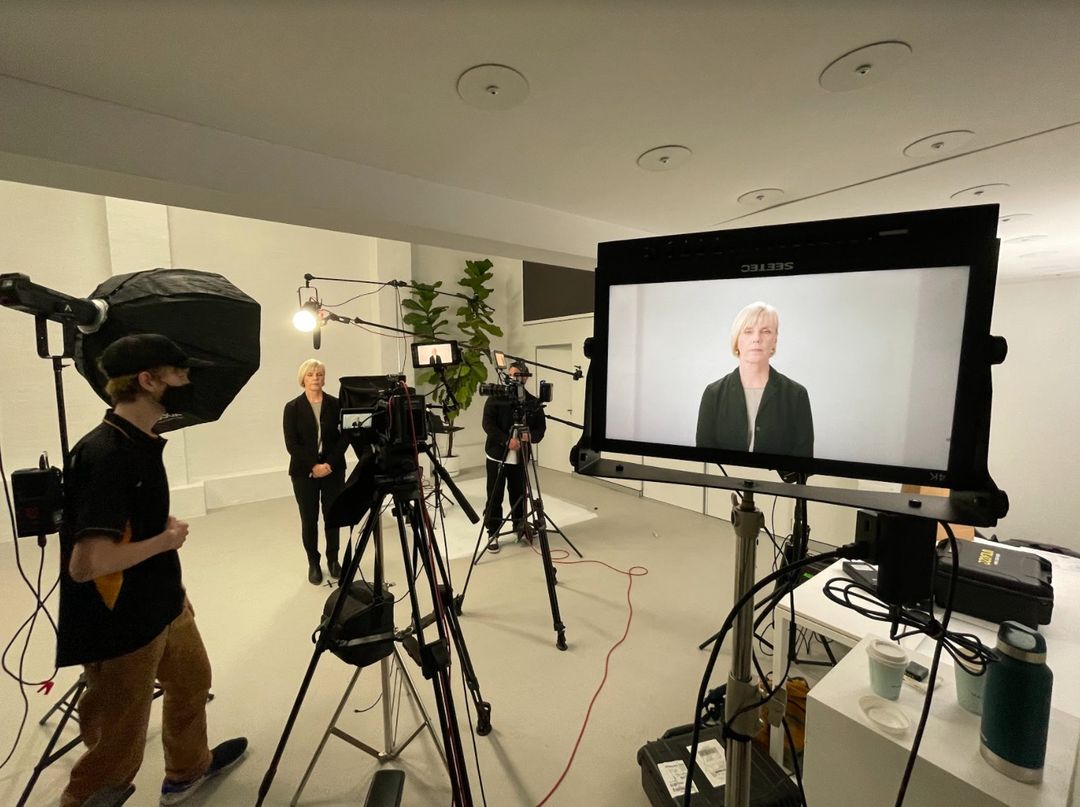 Behind-the-scenes film shoot features an adult woman being filmed by a camera crew in a brightly lit photo studio with plants.