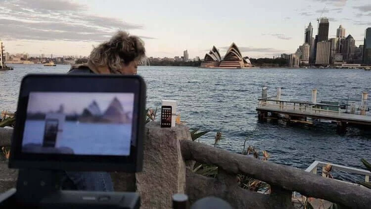 Sydney Opera House and city skyline backdrop a woman filming a cell phone on a stone railing by blue water, shot on camera display.