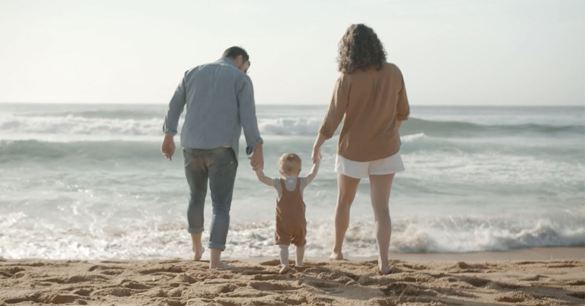 Back view of a family of three walking towards the ocean on a sandy beach. A man, woman, and child hold hands near ocean waves.