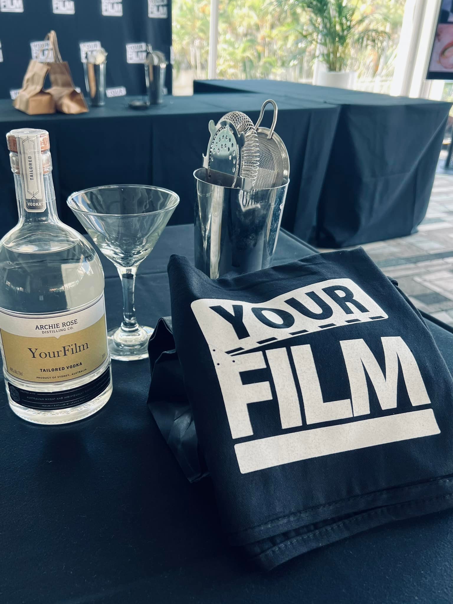 A table displays a bottle of liquor, martini glass, metal bar tools and a t-shirt with the words "Your Film" in an indoor setting.