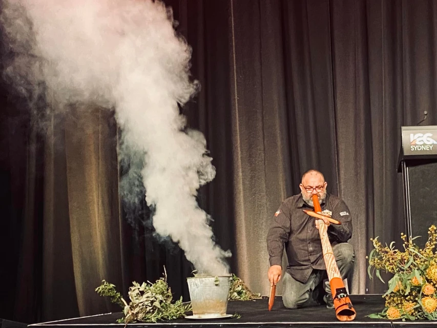 Adult male performing smoking ceremony with didgeridoo on stage, using native plants and a metal container. Smoke filling the scene.