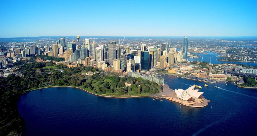High-angle view of Sydney, Australia, featuring the Opera House, a harbor with boats, bridges, and skyscrapers on a clear, sunny day.
