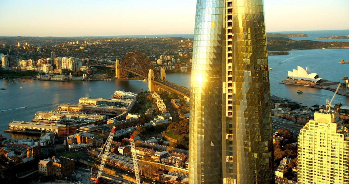 Aerial view of Sydney Harbour featuring the iconic Sydney Opera House and Harbour Bridge, alongside modern skyscrapers in golden light.