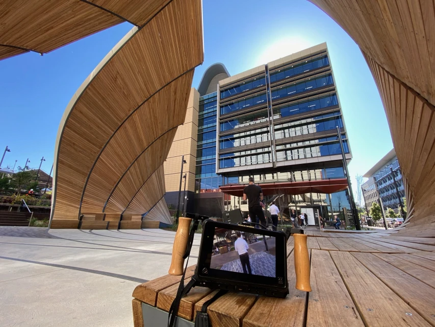 Outdoor scene captures a modern glass-clad building under a blue sky, framed by curved wooden structures, with film equipment in the foreground.