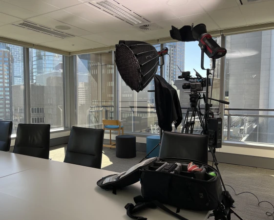 Camera and lighting equipment set up in a bright modern office interior with floor to ceiling windows and city view, ready for filming.