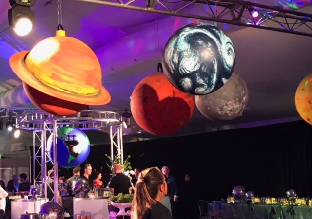 Event space with hanging planet decorations, people, tables with centerpieces, and bright event lighting with trusses overhead.