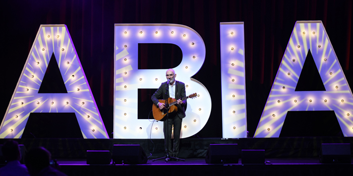 Man playing acoustic guitar on a stage with illuminated letter props behind him, wearing a dark suit. Concert setting with stage lights.