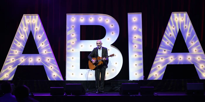 Man playing acoustic guitar on a stage with illuminated letter props behind him, wearing a dark suit. Concert setting with stage lights.