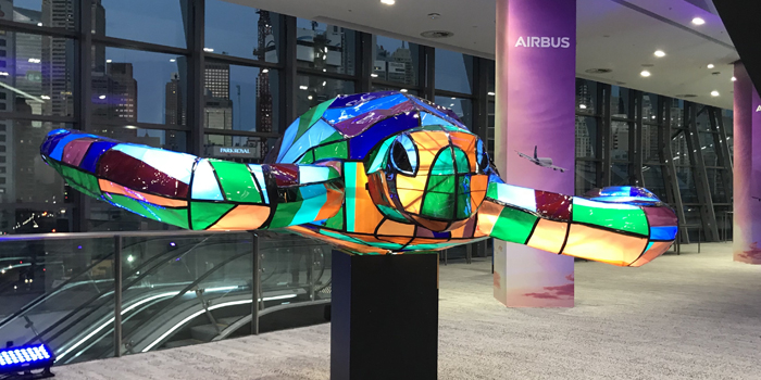 Colorful mosaic sea turtle sculpture on a black pedestal inside a building with large windows and a cityscape view in the background.