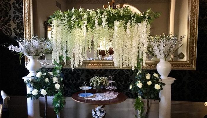 Interior with a table, floral arrangements, and a mirror. White wisteria and other flowers create an elegant, decorative display.