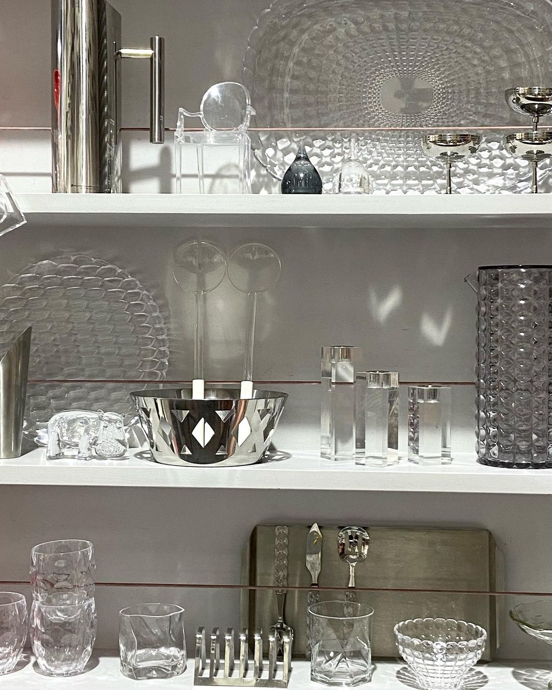 Still life arrangement on shelves featuring crystal glassware, metal pitchers, bowls, tableware, and decorative objects in an indoor setting.