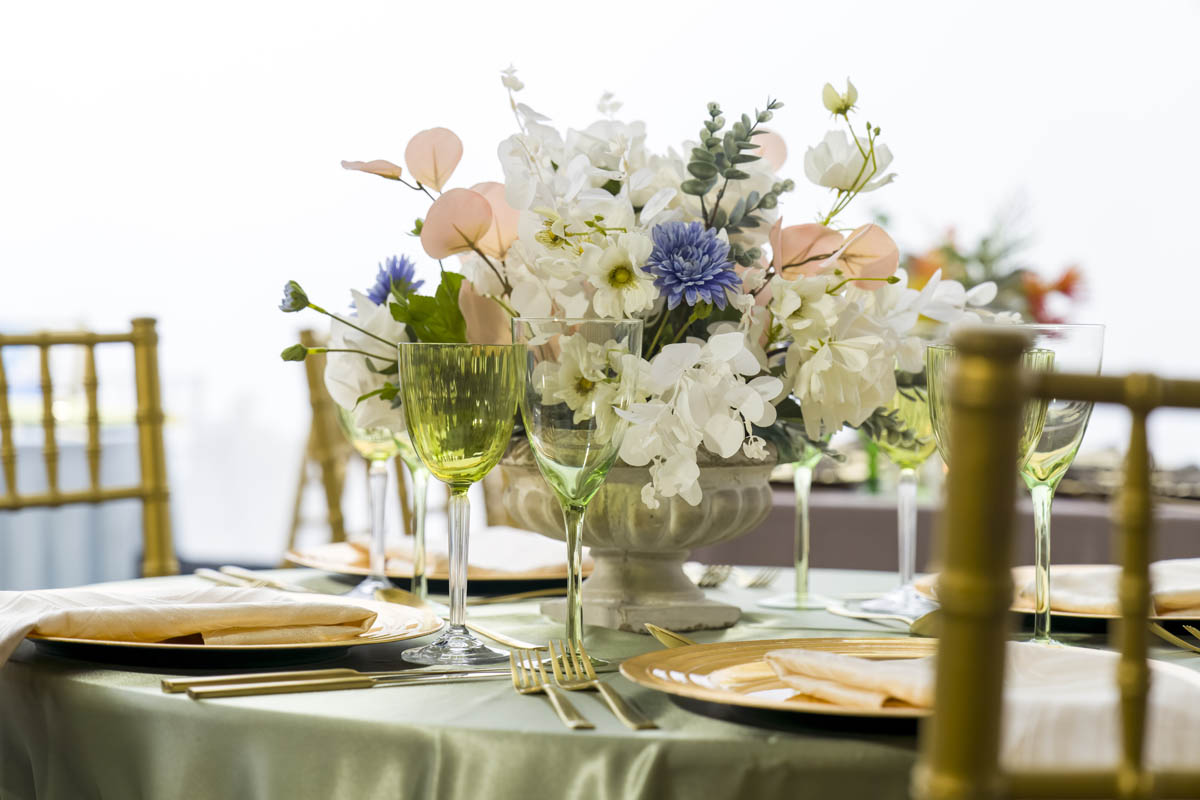 Elegant table setting with a floral centerpiece featuring white, blue, and peach flowers, complemented by green-tinted glassware and gold flatware.