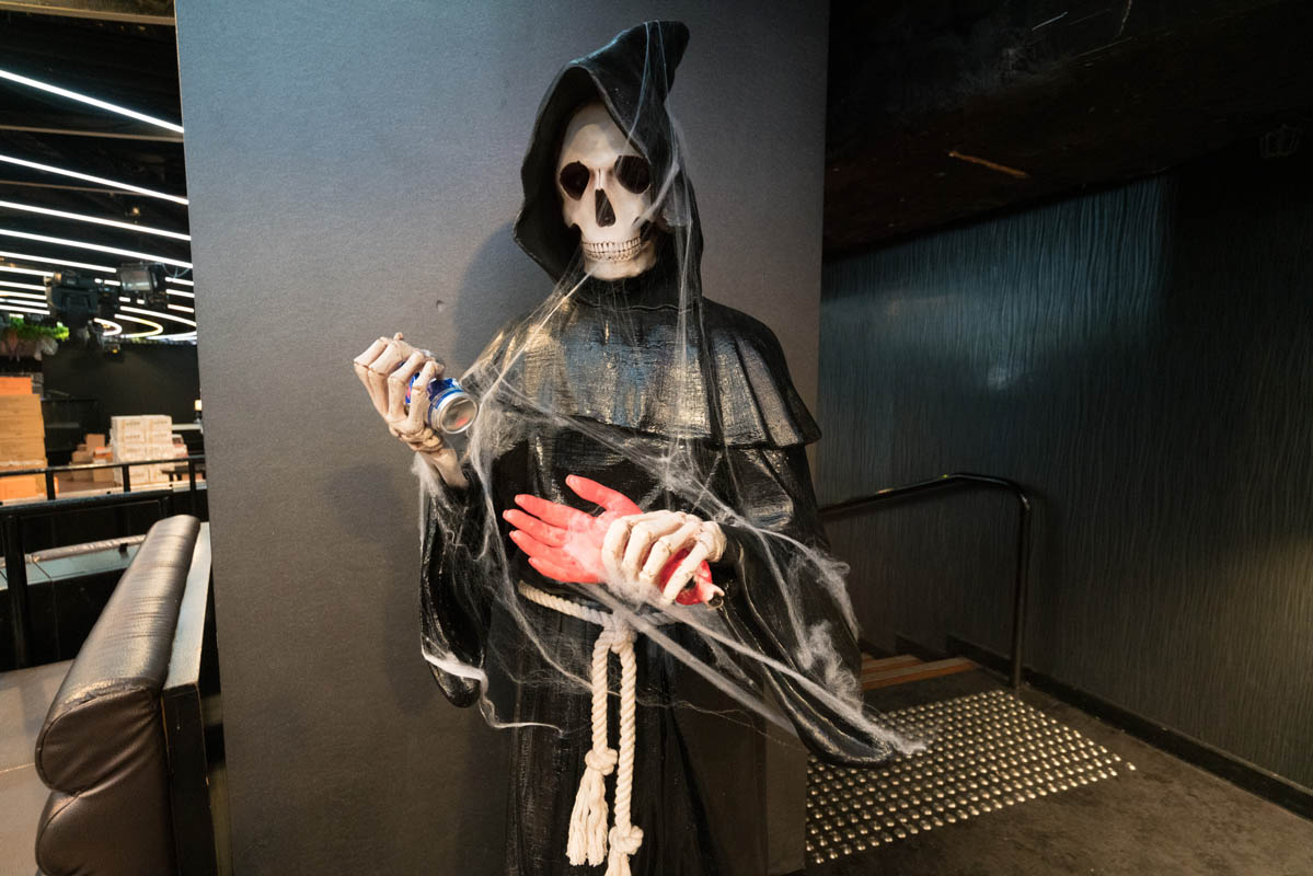 Grim reaper decoration with a can in a black hooded robe and fake spider webs in a dim interior space, Halloween or themed event.