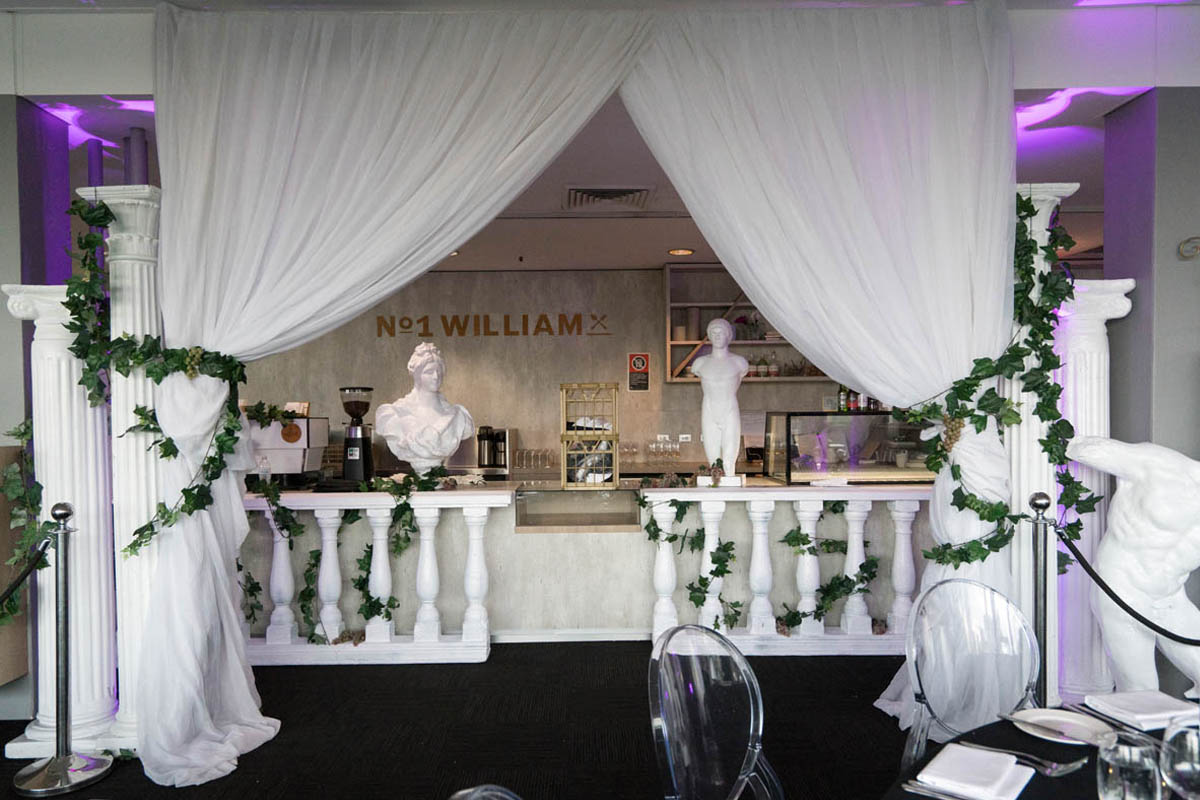 Elaborate interior design with white columns, draped fabric, classical busts, and green vines creating an elegant architectural display.