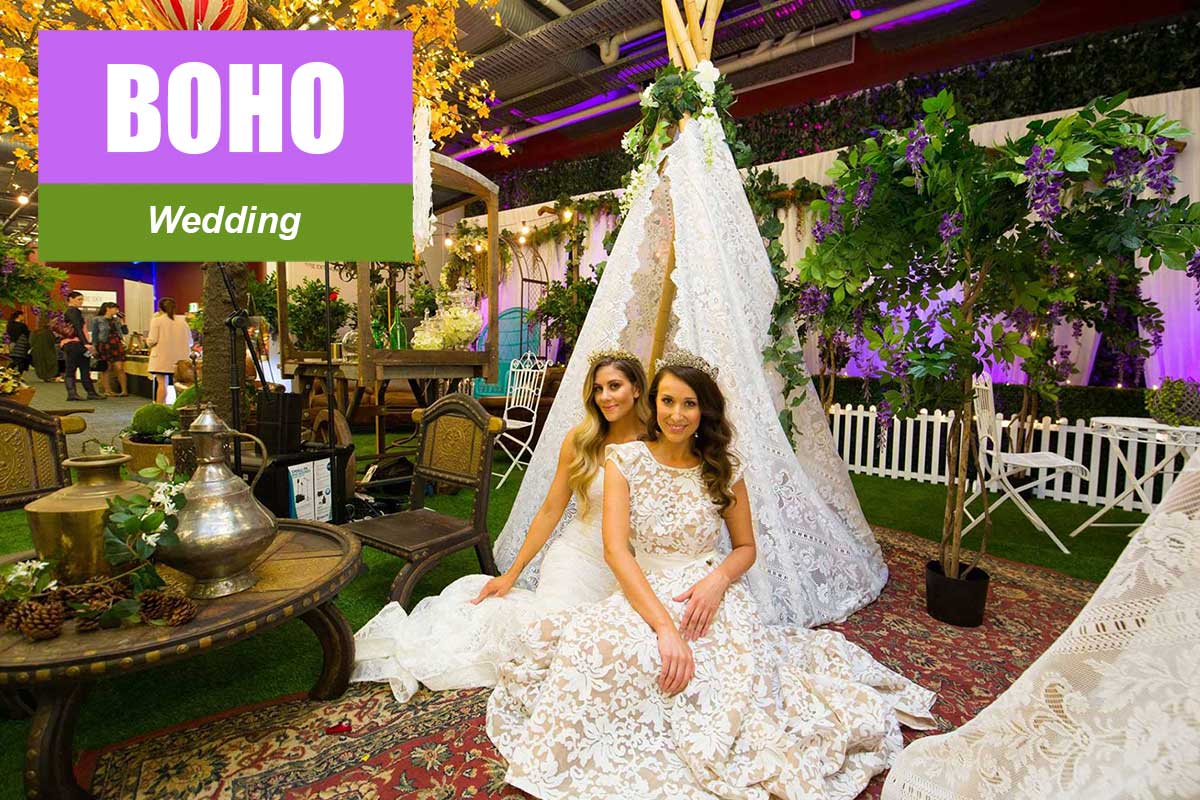 Two adult women in lace gowns pose in front of a lace teepee, indoor setting, with green and purple floral accents, and a vintage carpet.