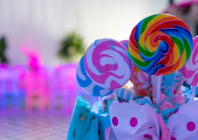 Colorful swirl lollipops on sticks are displayed in a decorative blue container with out-of-focus background of pink lights and greenery.