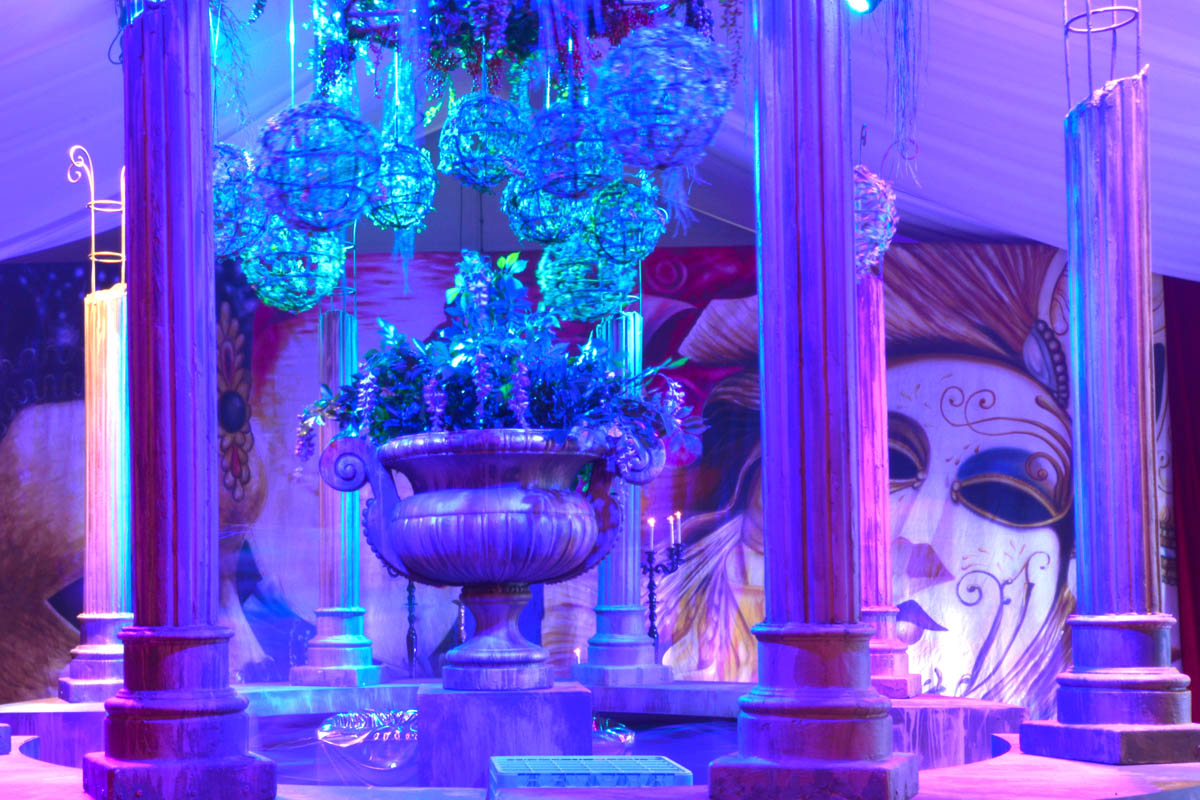 Ornate indoor display with an urn filled with greenery and decorative hanging spheres under blue lighting, set against painted backdrops.