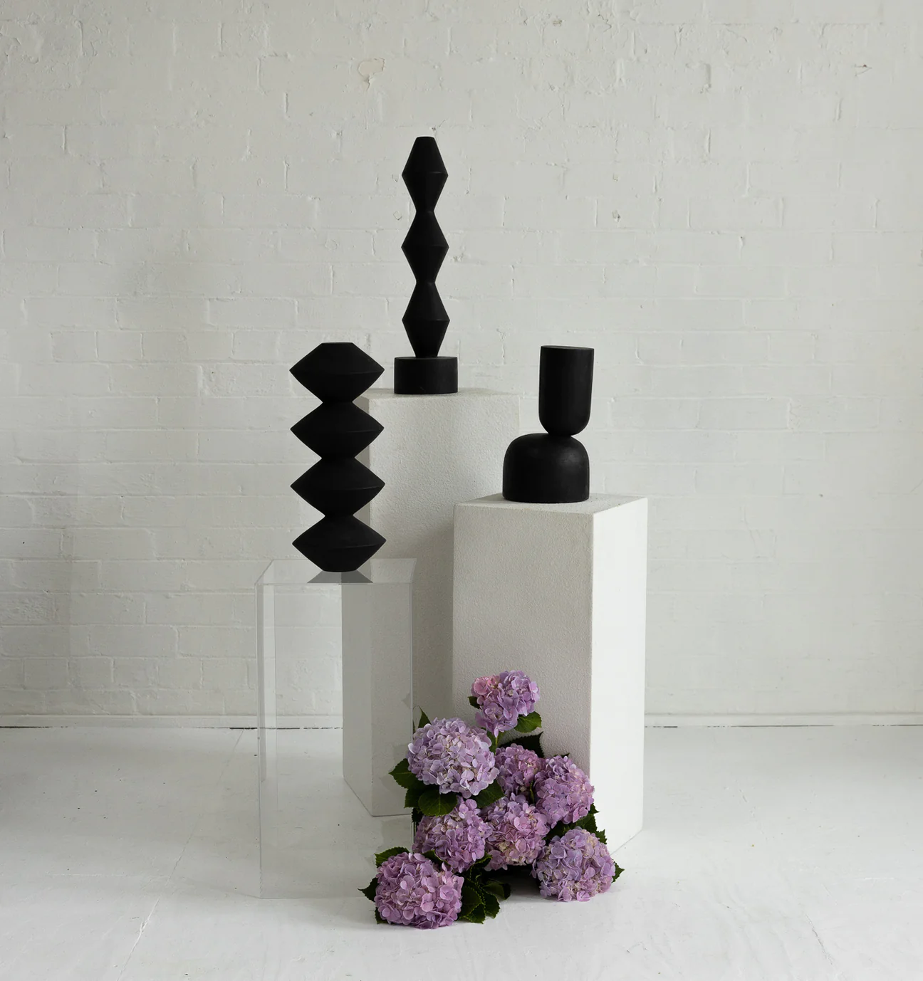 Three geometric, black ceramic vases are displayed on white plinths with light purple hydrangeas in a studio with a white brick wall.