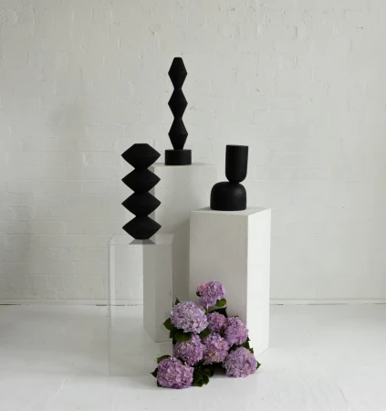 Three geometric, black ceramic vases are displayed on white plinths with light purple hydrangeas in a studio with a white brick wall.