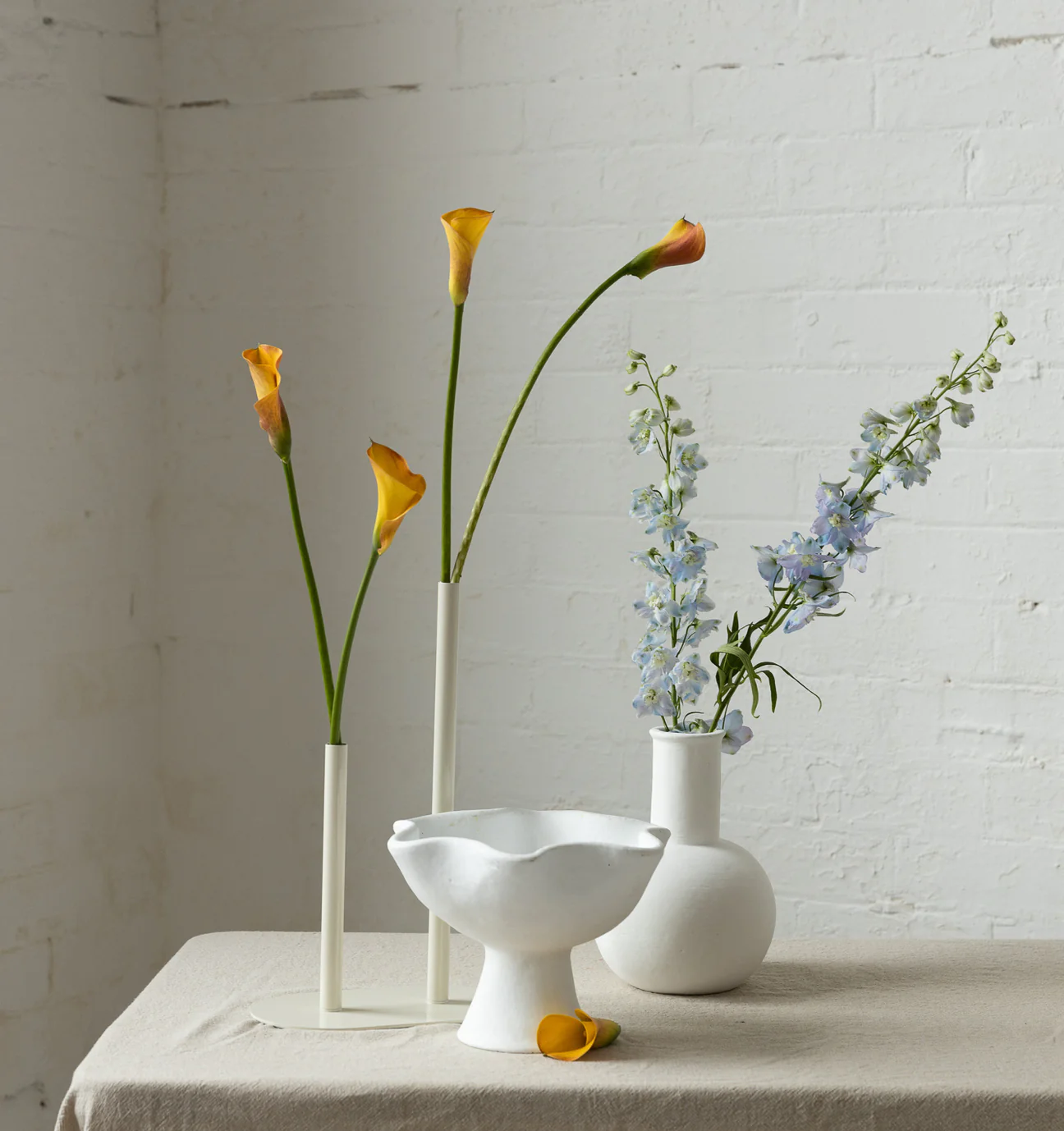 Still life arrangement of calla lilies in slender vases and light blue delphinium in a round white vase on a linen-covered table.