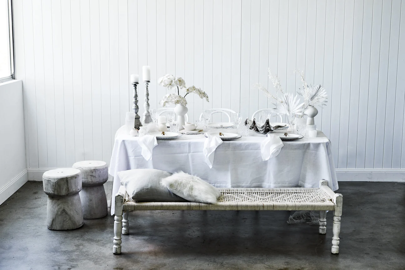 Elegant table setting with white linens, candles, flowers, and decorative objects, creating a serene and inviting atmosphere against a white backdrop.