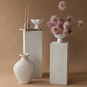 Still life shot of dahlia flowers in a white vase, white bowl and candles on pedestals, all against a tan backdrop.