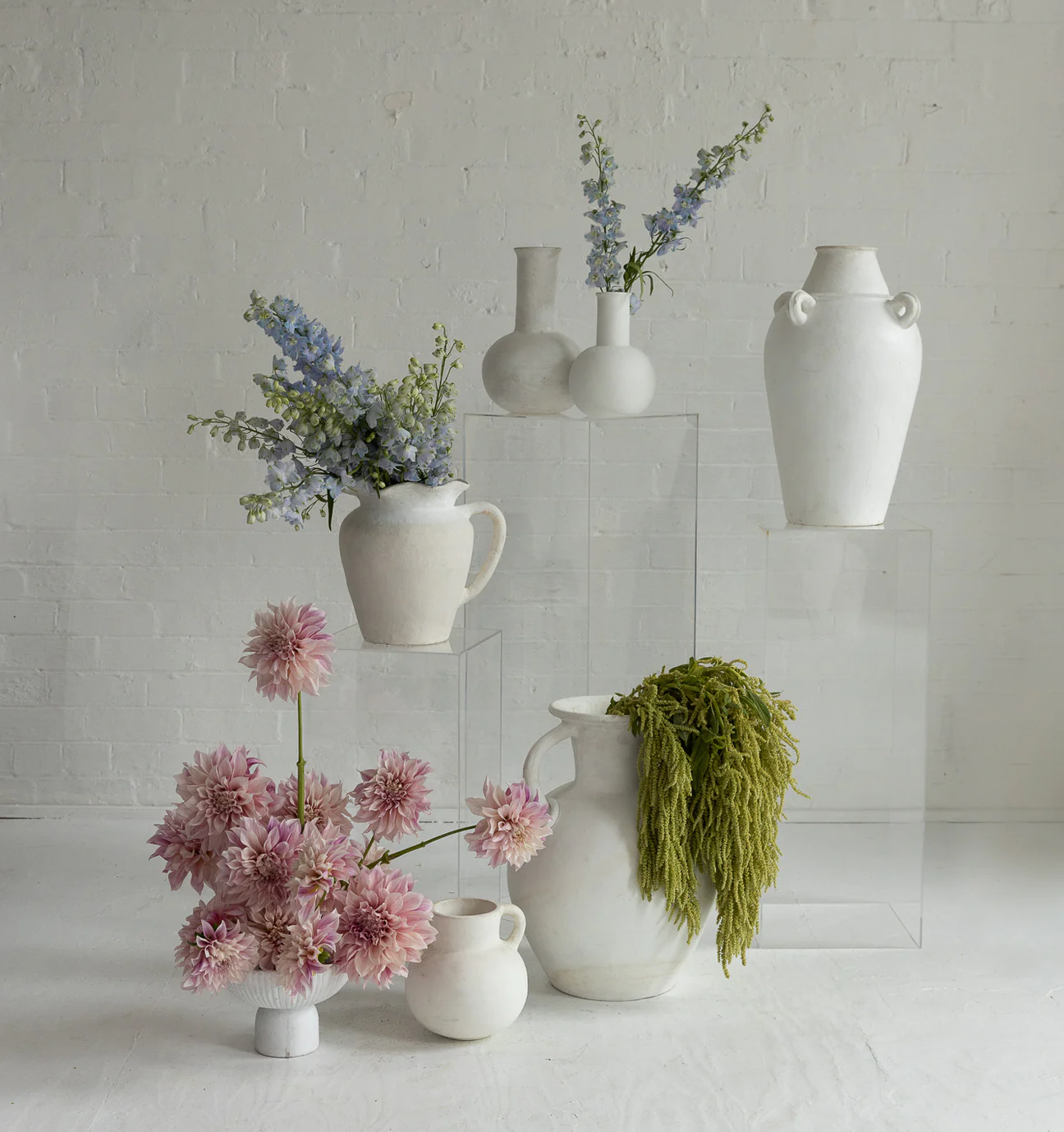 Still life of white ceramic vases of different shapes and sizes holding blue and pink flowers, displayed on clear acrylic pedestals.