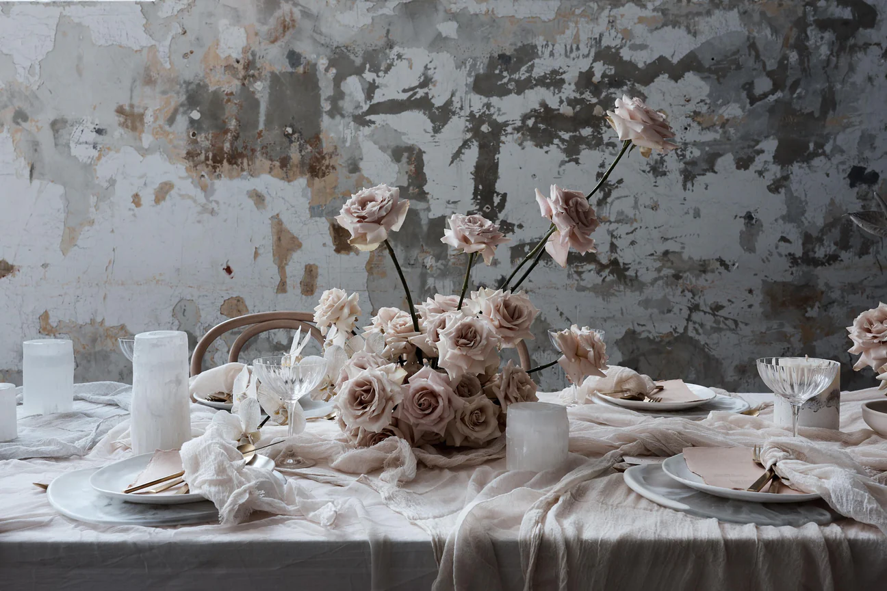 Floral centerpiece of pale roses on a set table with white linens, plates, glasses, and candles in an indoor space with a grey wall.