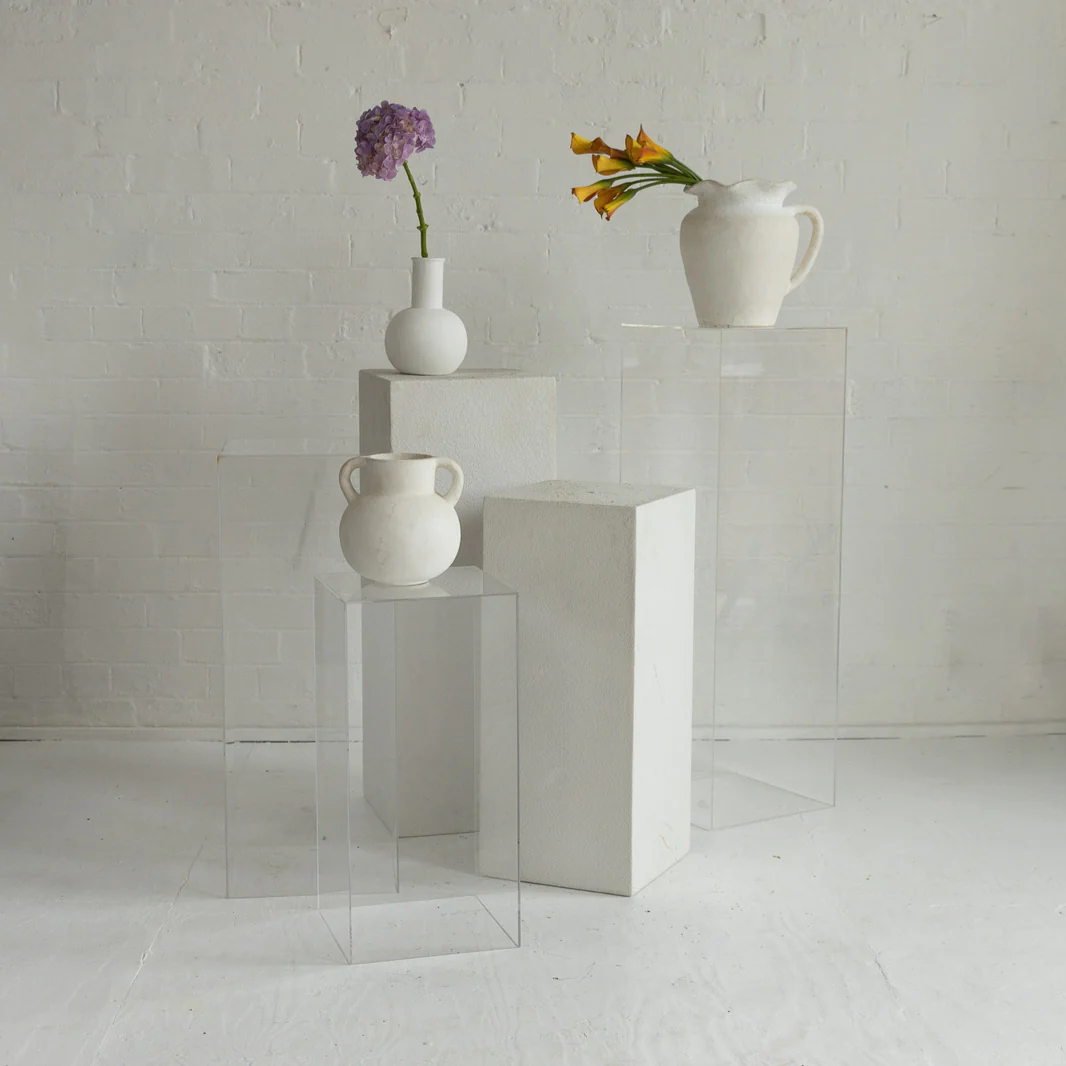 Still life of white vases with fresh flowers arranged on clear pedestals against a white brick wall in a minimalist aesthetic.