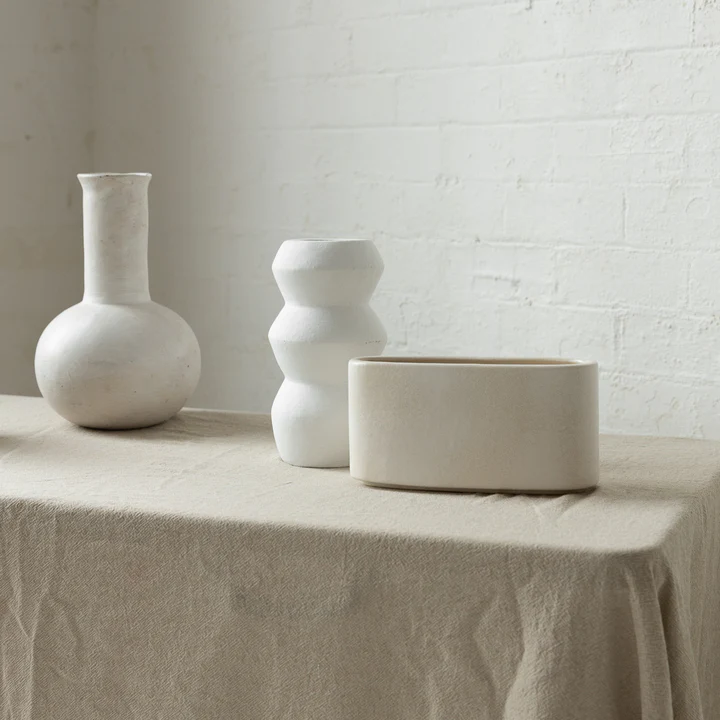 Three vases sit on a tablecloth in front of a white brick wall. The vases are white and cream coloured, and varying in shape.