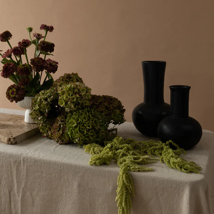 Still life with dark purple and green floral arrangements in vases, arranged on a table with a light neutral backdrop.